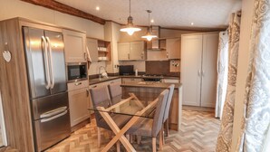 A modern kitchen with a stainless steel refrigerator, a built-in oven, and a microwave. There is a glass dining table with chairs. The kitchen has wooden cabinets and a herringbone floor pattern. - Avonal Lodge (Perth)