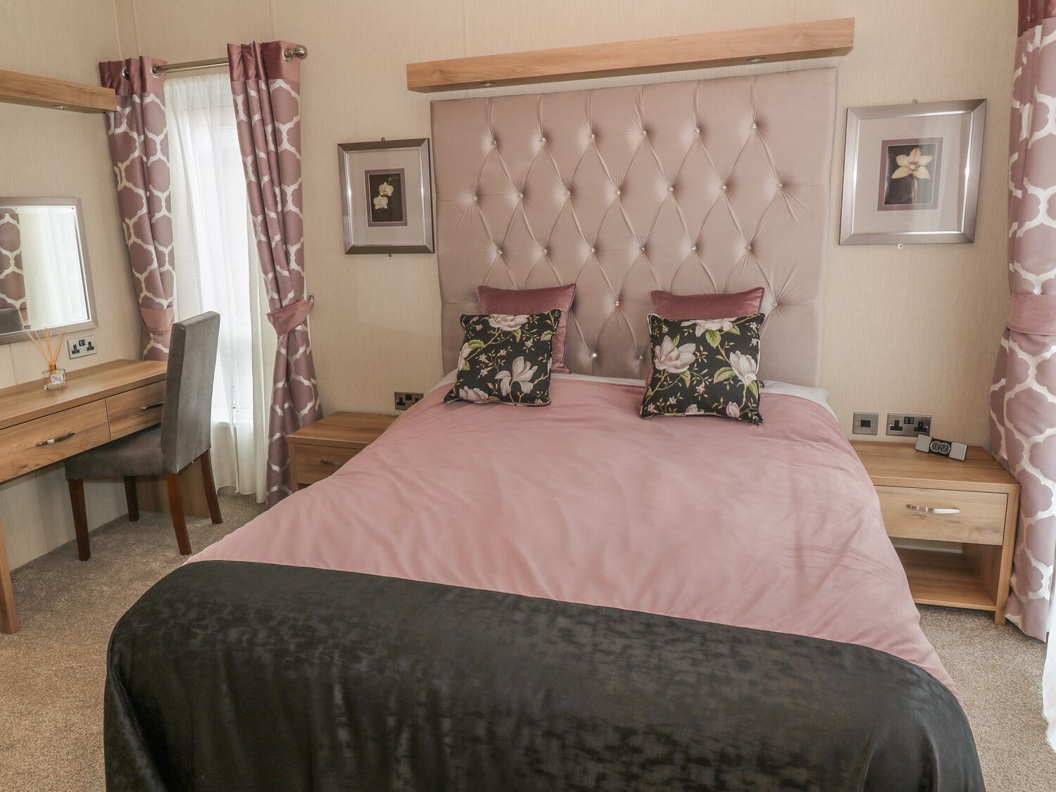 Quarto com cabeceira estofada, cama com roupa de cama rosa, duas almofadas florais, mesa de cabeceira de madeira com um telefone e uma escrivaninha com uma cadeira.