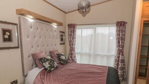 A bedroom with a tufted headboard, a wooden nightstand, a bed with a pink bedspread and floral pillows, a large window with sheer curtains, and a chandelier. - Avonal Lodge (Perth)