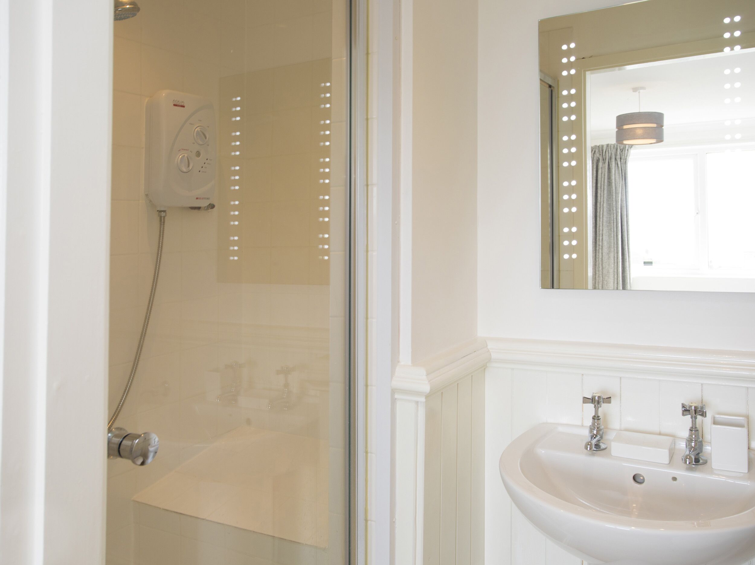 A bathroom with a glass shower enclosure, a white sink with two faucets, and a mirror with lights.