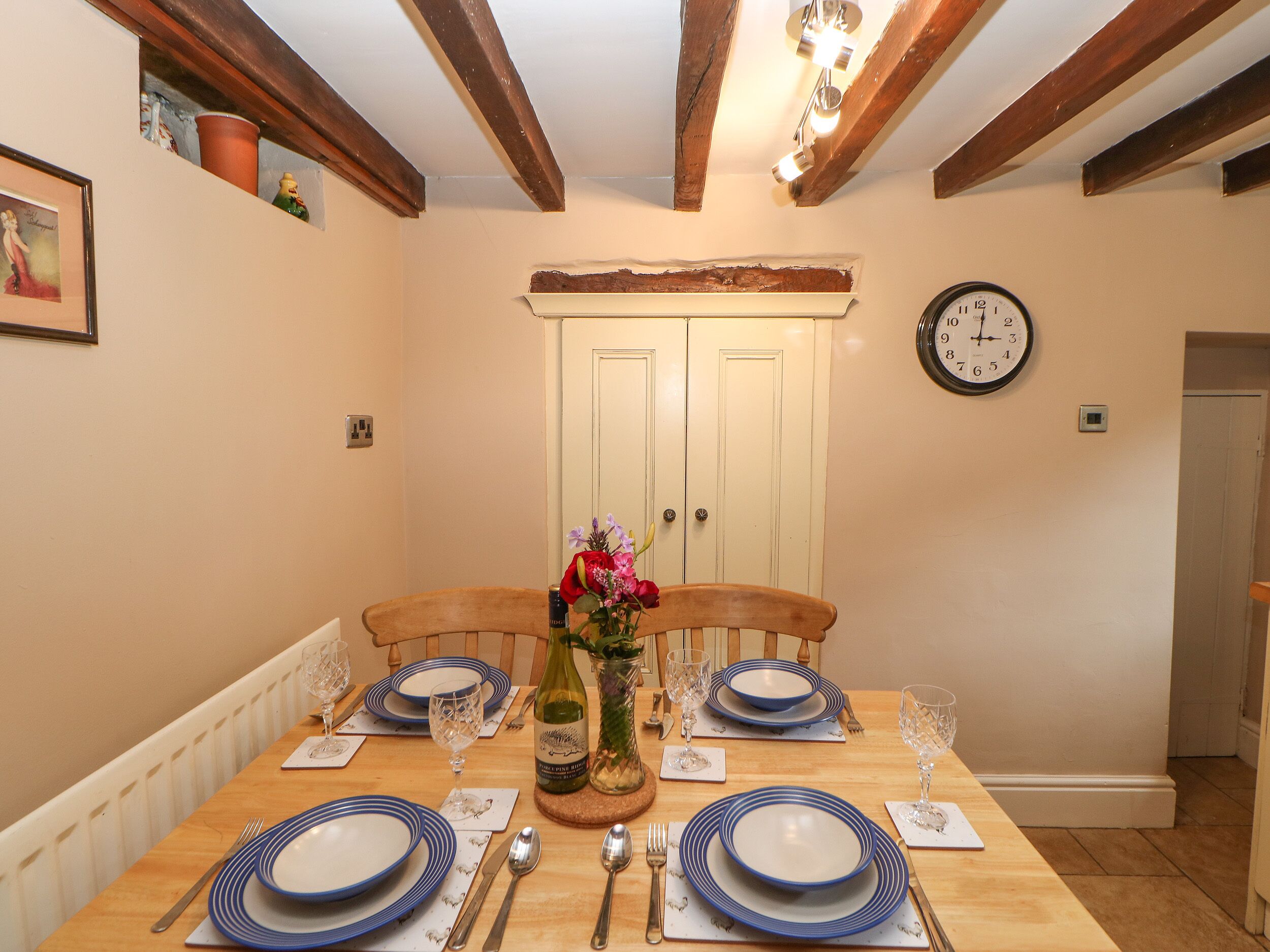 Comedor con una mesa de madera dispuesta para cuatro, un florero con flores y un reloj en la pared.