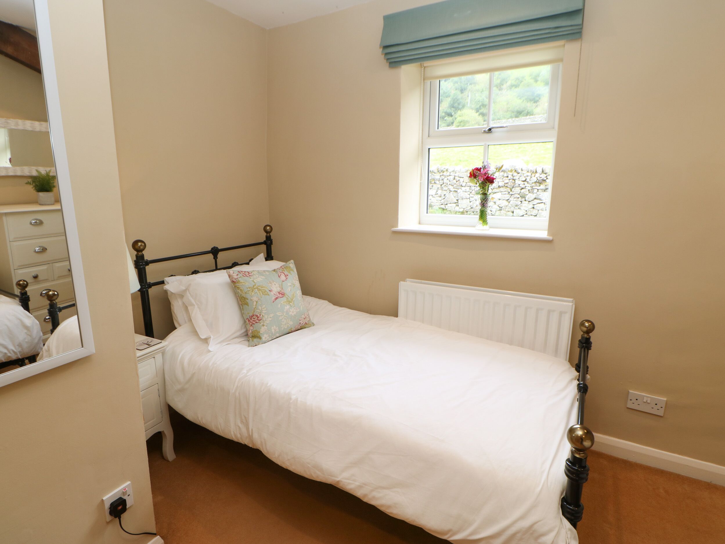A neatly made bed with a floral pillow, a dresser with drawers, a mirror, a window with a view of greenery, and a radiator.