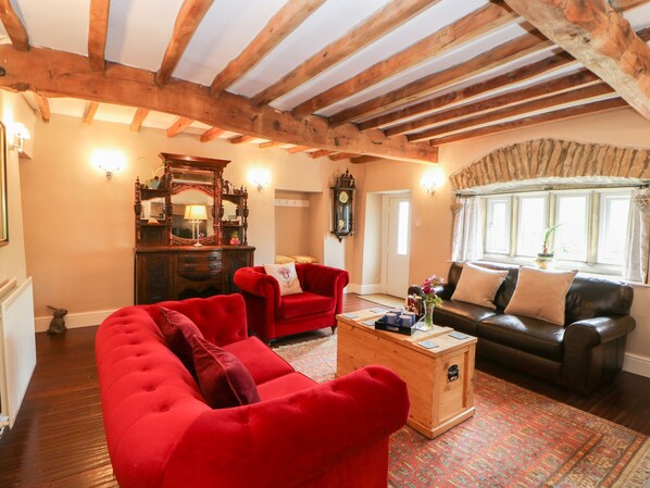 A cozy living room with a red sofa, a black leather sofa, and a wooden coffee table. There is a wooden cabinet with a mirror, a clock, and a lamp. The room has wooden beams on the ceiling and a window with curtains. - Cowlings (Richmond)