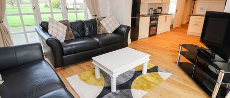 A modern living room with a black leather sofa, a white coffee table, and a TV. The room has wooden flooring and a view of a garden through large windows.
