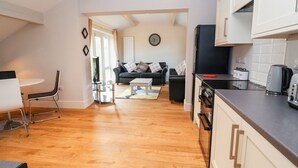 A modern kitchen with white cabinets, a black refrigerator, and a coffee maker. There is a dining area with a white table and chairs, and a living room with a black sofa and a coffee table. - Commonwood Cottage (Wrexham)