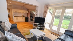 A modern living room with a black leather sofa, a white coffee table, and a TV on a stand. There is a kitchen in the background with white cabinets and a brick accent wall. - Commonwood Cottage (Wrexham)