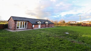 A brick house with a sloped roof, a fenced yard, and a grassy field. - Commonwood Cottage (Wrexham)
