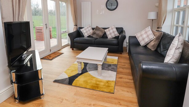 A living room with a black leather sofa, a white coffee table, a black TV stand, and a patterned rug. - Commonwood Cottage (Wrexham)