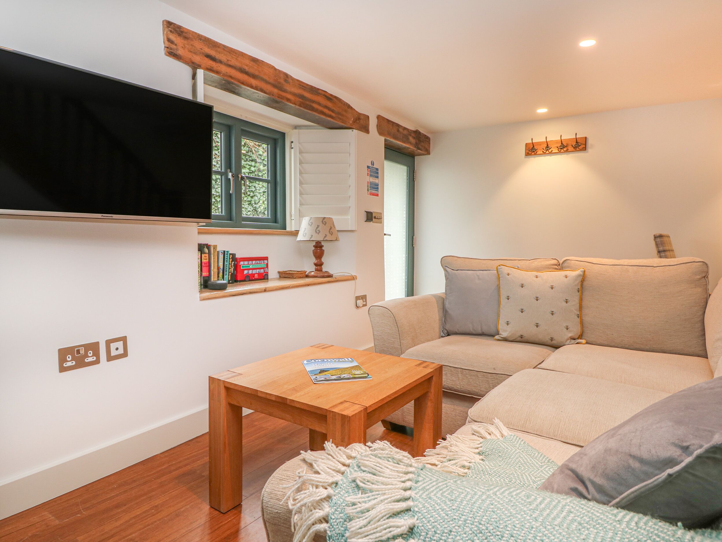 Una sala de estar acogedora con una mesa de madera, un sofá con cojines, un televisor montado y una ventana con contraventanas.