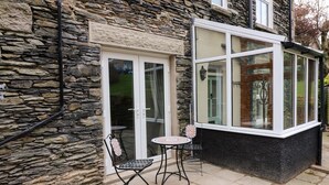 A stone building with a glass door and windows, a small patio with two chairs and a table, and a stone wall. - Giddy Goose (Windermere)