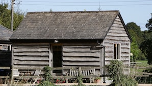 Eine rustikale Holzhütte mit geneigtem Dach, einer Veranda mit Bänken und einem kleinen Gartenbereich mit Blumen und Sträuchern.