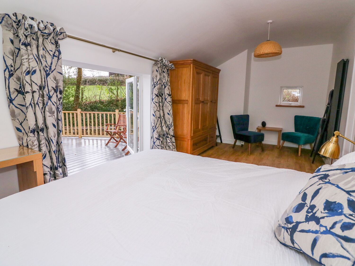 A bedroom with a large bed, a wooden wardrobe, a small table, two chairs, and a balcony with a view of greenery.