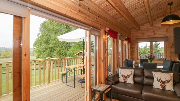 A wooden cabin with a deck, a sofa with deer-themed pillows, and a dining area with chairs and a table. - Sunnyside Lodge (Crewkerne)