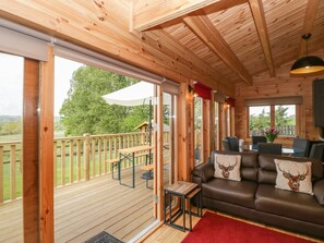 A wooden cabin with a deck, a sofa with deer-themed pillows, and a dining area with chairs and a table. - Sunnyside Lodge (Crewkerne)
