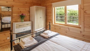 A wooden cabin bedroom with a bed, a wardrobe, a dresser, a mirror, and a window with a view of greenery. - Sunnyside Lodge (Crewkerne)