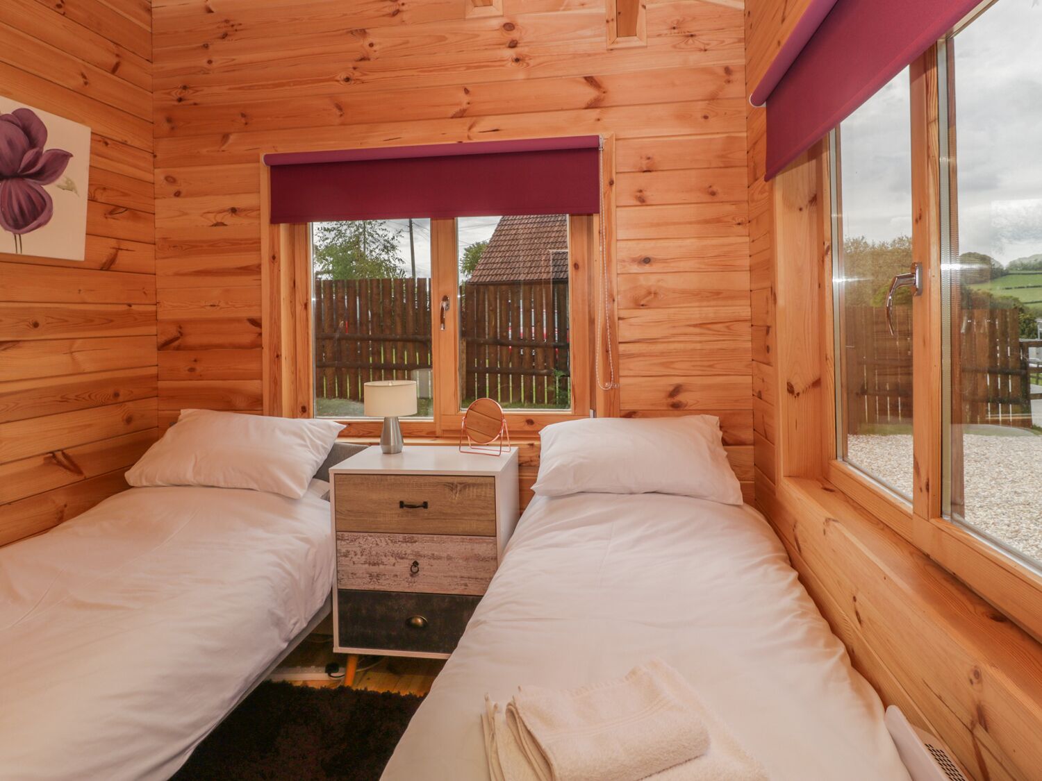 A wooden cabin room with two beds, a nightstand, a window with a view of a wooden fence and a house, and a purple curtain.