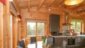 A wooden cabin interior with a glass dining table, chairs, a kitchen with modern appliances, and a large window. - Sunnyside Lodge (Crewkerne)