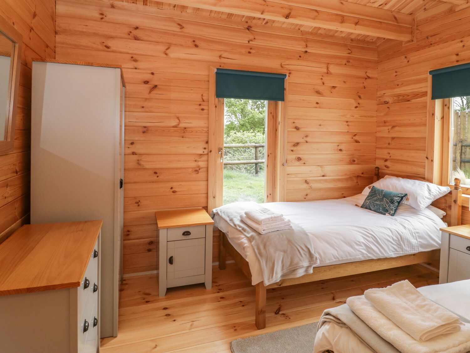 Un dormitorio de cabaña de madera con cama, mesitas de noche, un pequeño armario y una ventana con vistas a la vegetación.