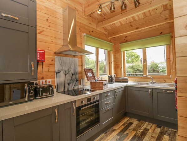 Une cuisine moderne avec des meubles gris, une hotte en acier inoxydable, un four à micro-ondes et un four traditionnel. On y trouve des fenêtres avec des stores verts, un plancher en bois et une vue sur la verdure à l’extérieur.
