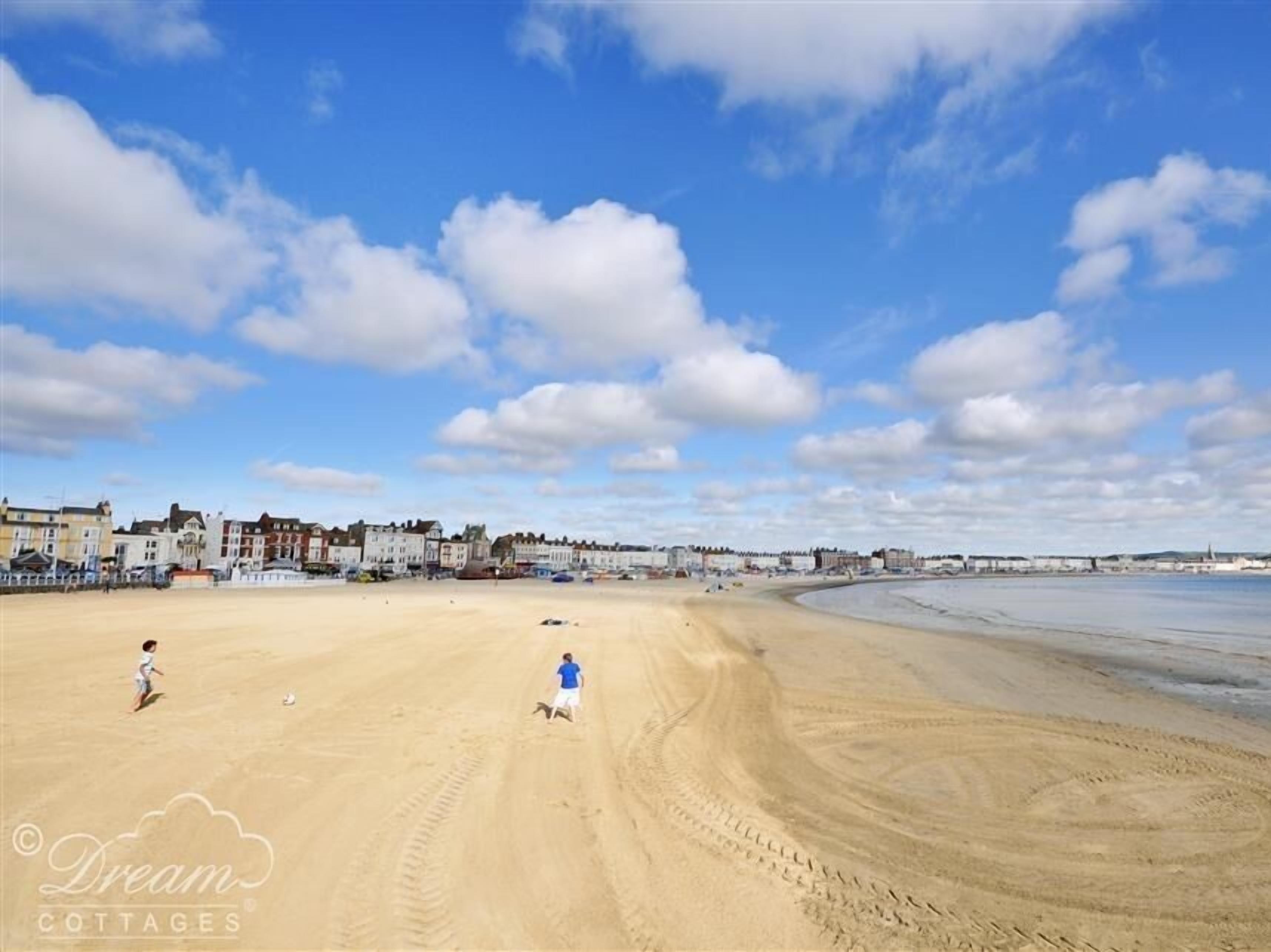 A sandy beach with footprints, a child, and a person running. There are buildings in the background.