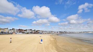 A sandy beach with footprints, a child, and a person running. There are buildings in the background.