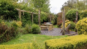 Um jardim com pérgula de madeira, mesa para piquenique e churrasqueira.