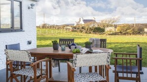 A wooden table with chairs, a potted plant, and a teapot on a patio with a view of houses and a field. - Mwythus (Holyhead)