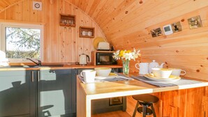 A cozy cabin interior with a wooden interior, a kitchen area with a sink, stove, and microwave, a dining table with cups and a vase of flowers, and a window providing natural light. - Chapel Pod (Saltash)
