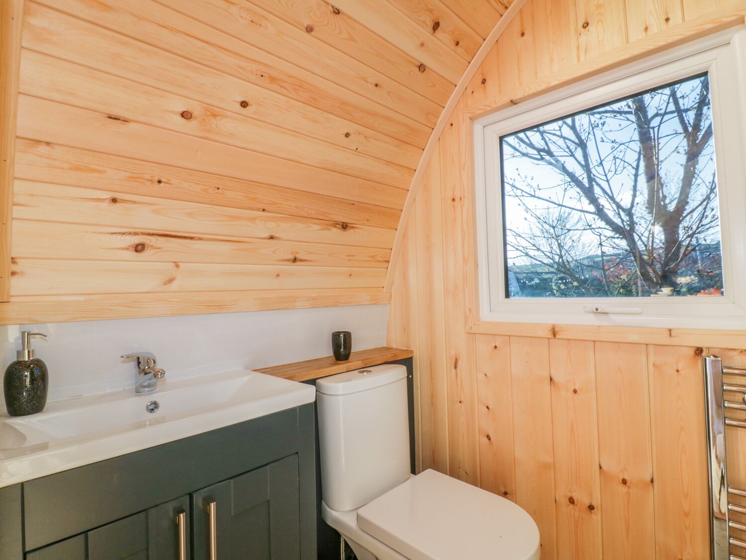 Une petite cabane en bois comprenant un coin cuisine équipé d’un évier, de toilettes et d’une fenêtre donnant sur la forêt.