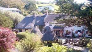 A traditional stone house with a thatched roof, surrounded by lush greenery and a wooden fence. - Warmwell Lodge (Dorchester)