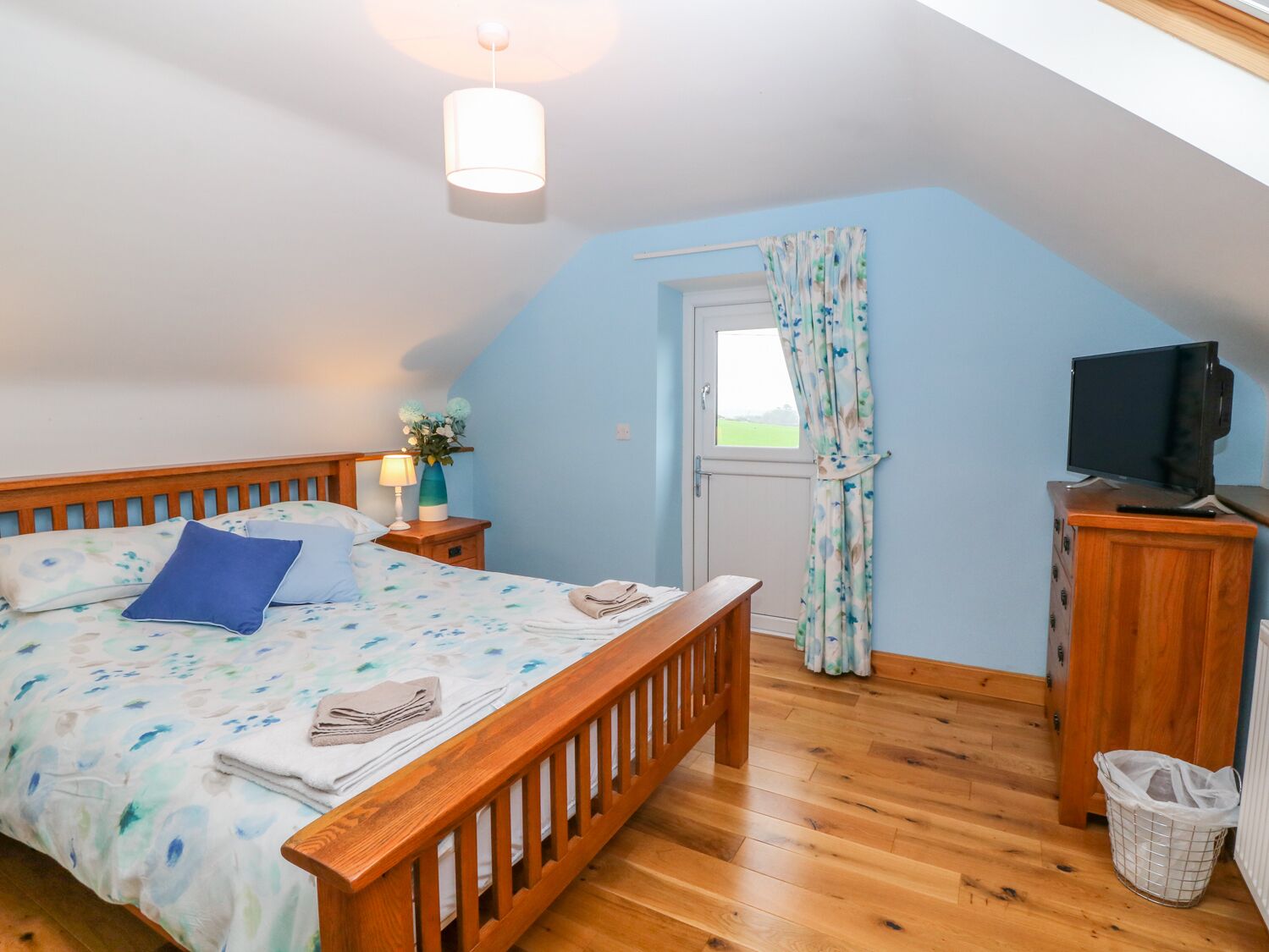 A bedroom with a wooden bed, a nightstand with a lamp, a television on a wooden cabinet, and a window with curtains.