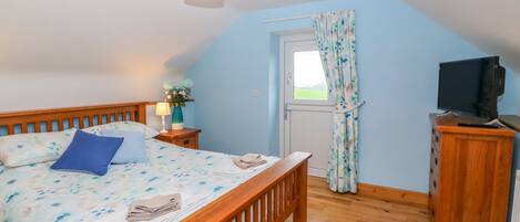 A bedroom with a wooden bed, a nightstand with a lamp, a television on a wooden cabinet, and a window with curtains.