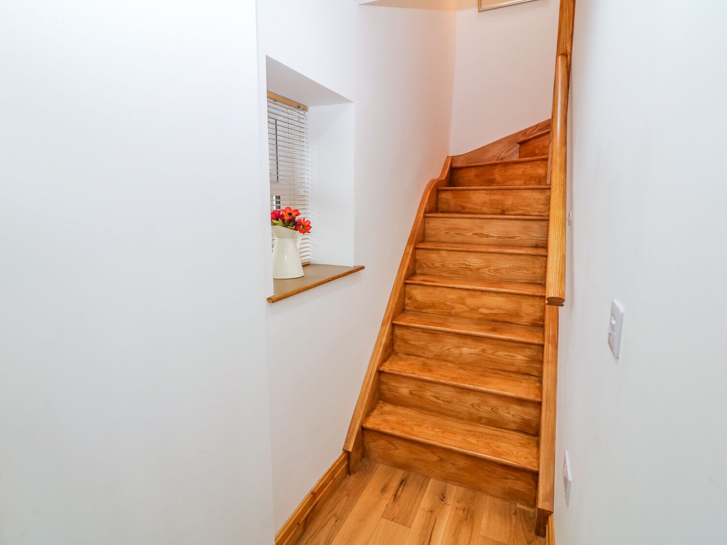 A wooden staircase in a hallway with white walls and a small window with a white vase of flowers.
