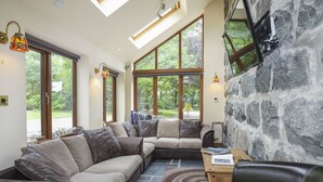 Uma sala de estar moderna com sofá seccional, poltrona de couro e parede com detalhes em pedra. Grandes janelas oferecem luz natural e vista para o exterior.
