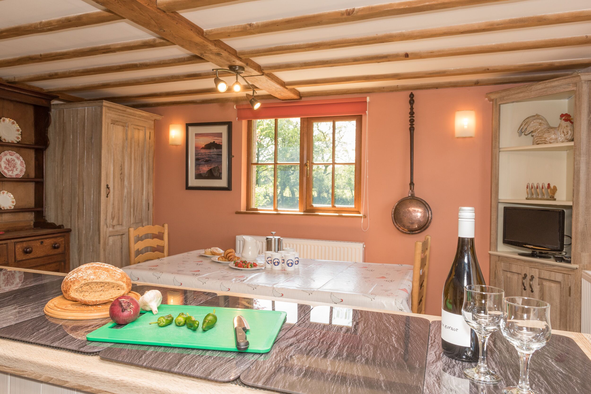 A kitchen with a wooden ceiling, a window with a view of greenery, a table with breakfast items, and a wine bottle.