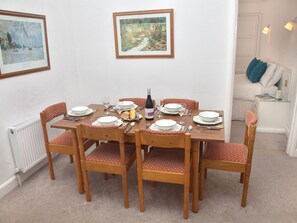 En spisesal med et træbord dækket til seks personer, omgivet af stole med mønstrede hynder. Væggene er pyntet med indrammede kunstværker, og i baggrunden ses en radiator og en hvid dør.