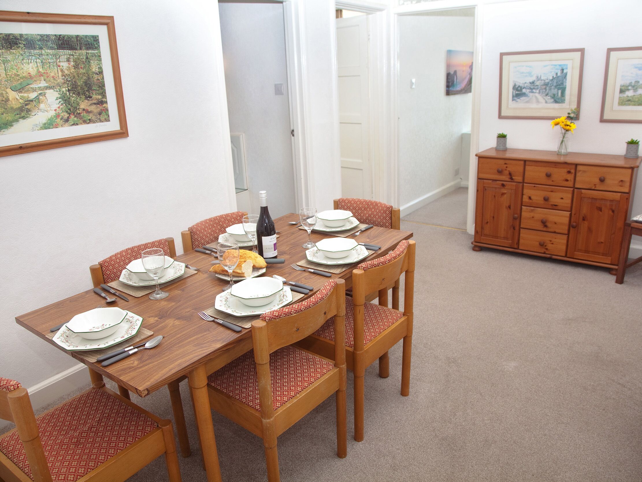Comedor con una mesa de madera dispuesta para cuatro, rodeada de sillas con cojines estampados. Hay una botella de vino y pan sobre la mesa.