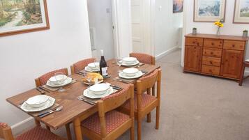 Comedor con una mesa de madera dispuesta para cuatro, rodeada de sillas con cojines estampados. Hay una botella de vino y pan sobre la mesa.