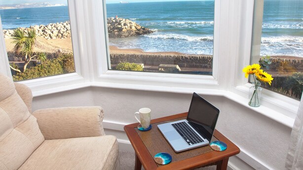 A room with a large bay window overlooking the ocean, a laptop on a small table, and a vase with yellow flowers. - Weymouth Bay Apartment B (Weymouth)