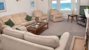 A living room with a beige sofa, armchairs, a coffee table, and a TV stand with a flat-screen TV and a small table with a vase. - Weymouth Bay Apartment B (Weymouth)