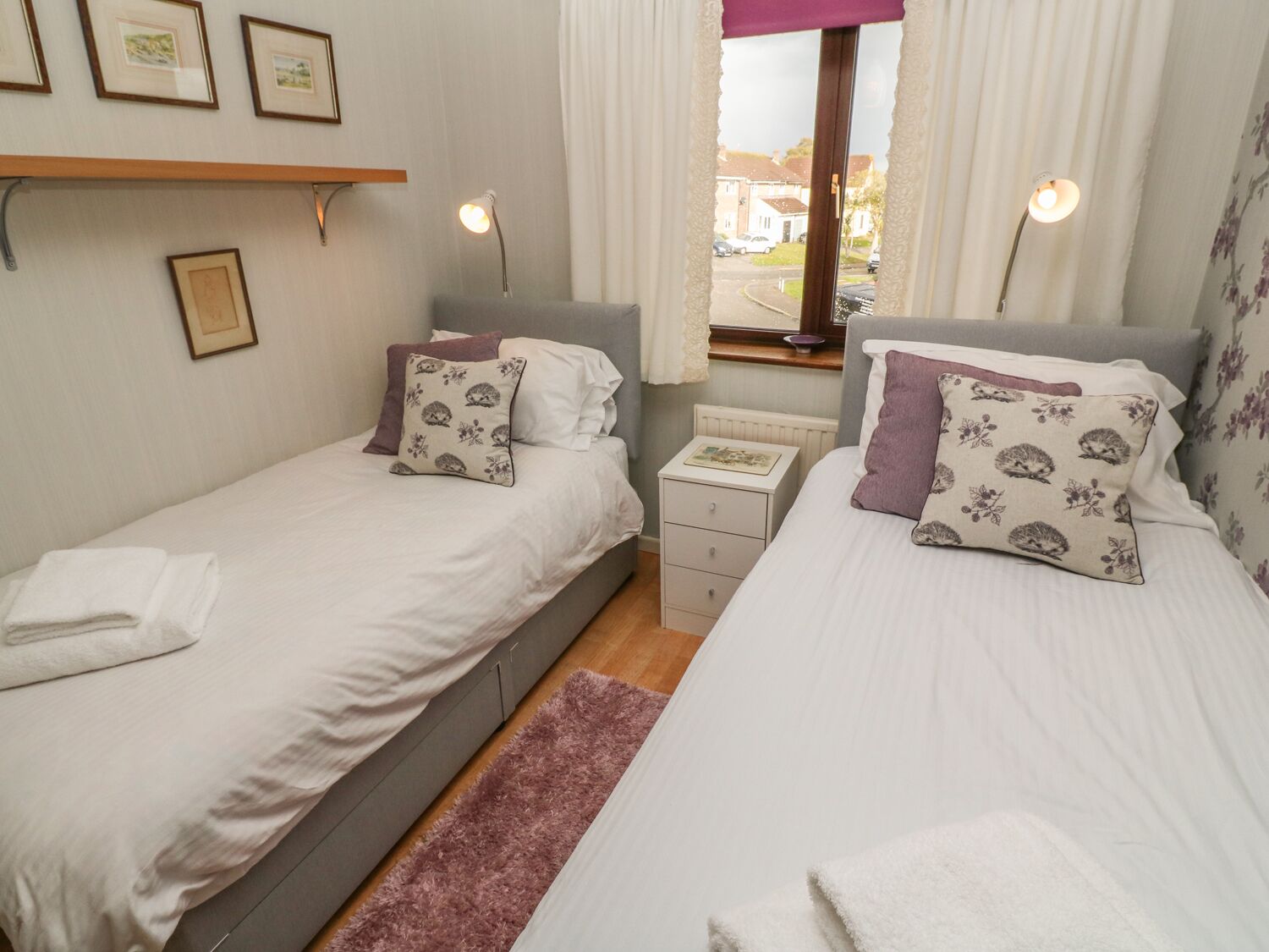 Un dormitorio doble con dos camas, una mesita de noche, una ventana con cortinas y cuadros enmarcados en la pared.