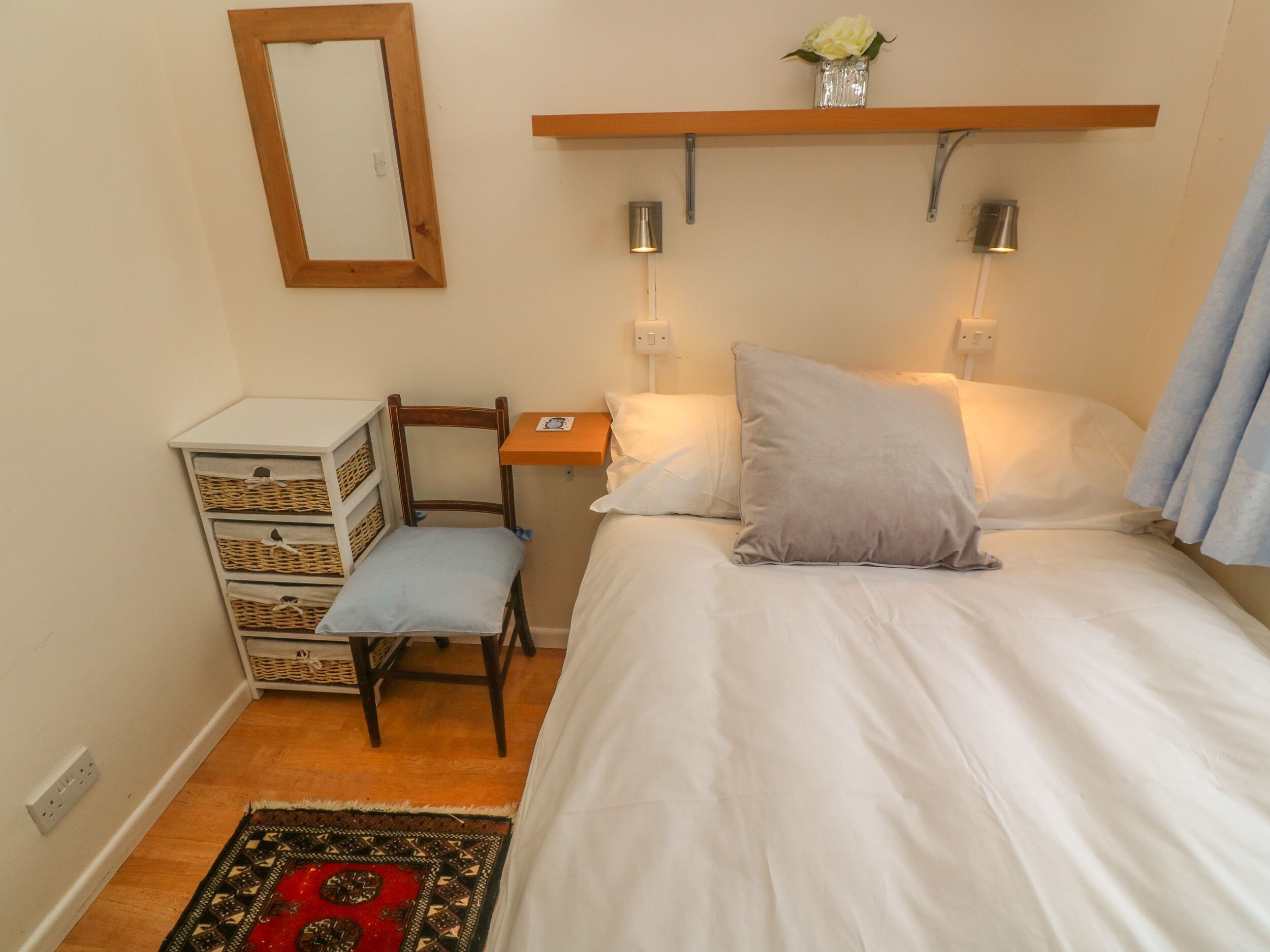 A small, neatly arranged bedroom with a single bed, a chair, a nightstand, a mirror, and a shelf with a vase.