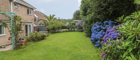 A residential garden with a lawn, flowering plants, and a brick house.