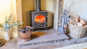 A stone fireplace with a wood-burning stove, a copper bucket of wood, and a wicker basket of logs. - Rose Cottage (Skipton)