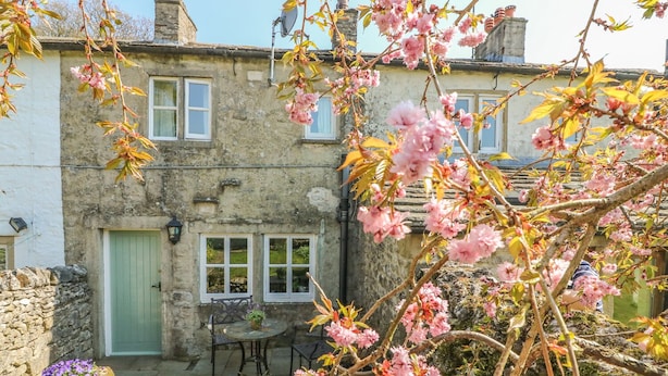 A stone house with a green door, windows, and a small patio with a table and chairs. - Rose Cottage (Skipton)