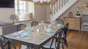 Comedor con una mesa de madera dispuesta para seis personas, rodeada de sillas negras. Un candelabro cuelga del techo, y se ve una escalera con barandilla blanca al fondo.