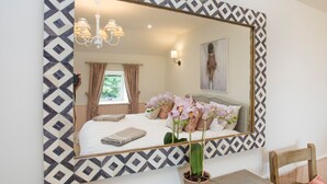 Habitación de hotel con un espejo grande, una cama con almohadas, una mesita de noche con un florero y una ventana con cortinas.