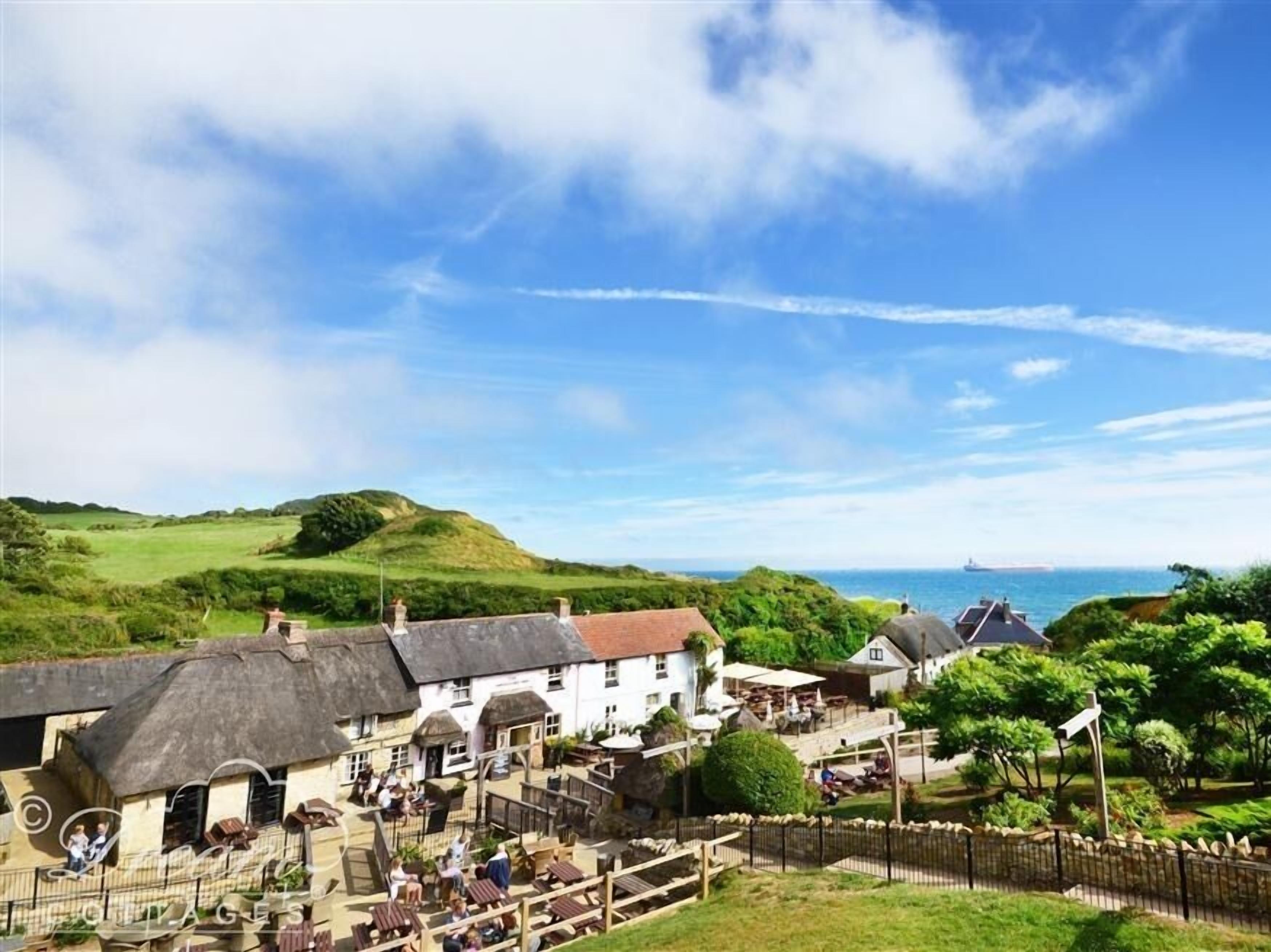 A coastal village with thatched-roof buildings, a central square with outdoor seating, and a view of the sea and hills in the background.