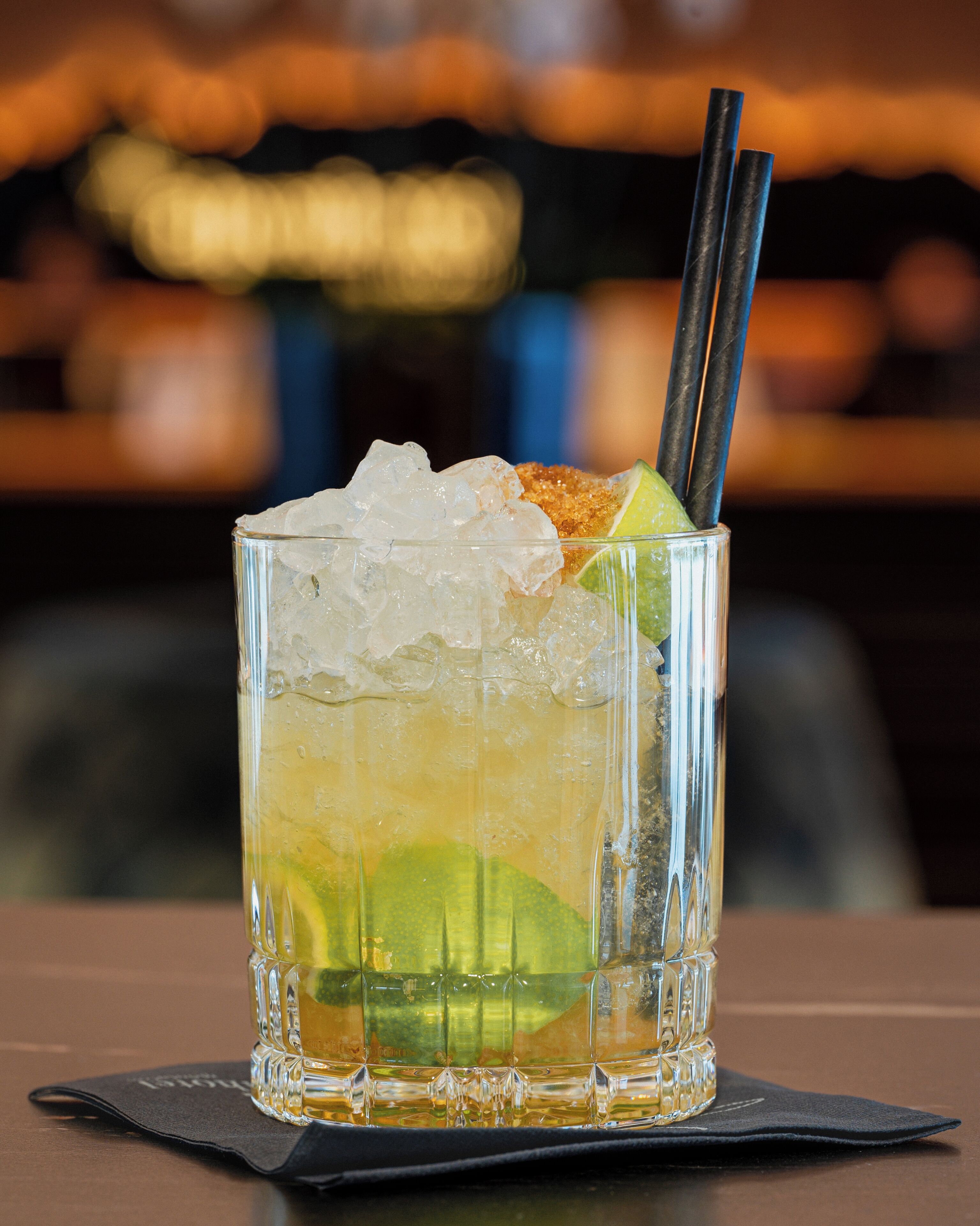 A glass of cocktail with ice cubes and a lime wedge, placed on a napkin on a table.