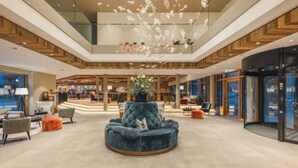 A spacious lobby with a central seating area, a high ceiling with a wooden panel design, and a view of the interior through large glass doors. - Alpenhotel Montafon & SPA (Schruns)
